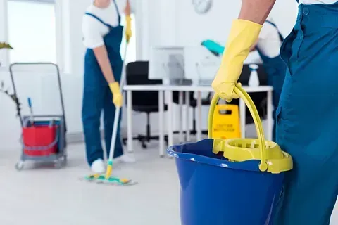 Personal de limpieza con monos azules y guantes amarillos. Uno sostiene un cubo, otros friegan una oficina blanca.