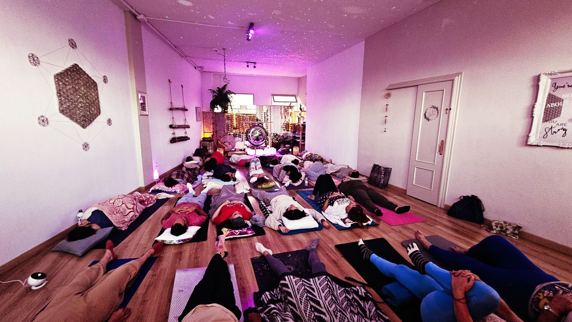 Personas tumbadas sobre colchonetas en un estudio de yoga. Suelo de madera, paredes blancas e iluminación violeta.