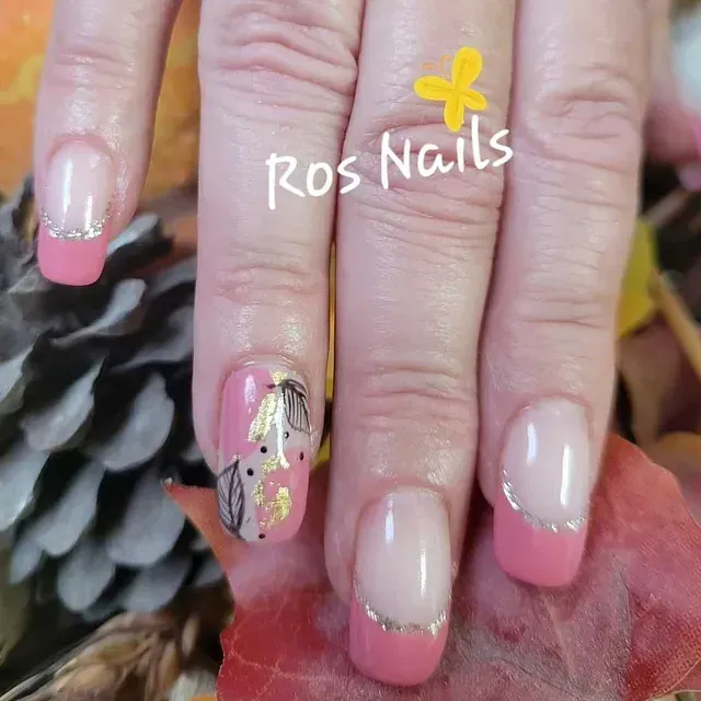 Una mano con un diseño de uñas rosa y dorado. Puntas francesas con detalles plateados y una uña con hojas y copos dorados.