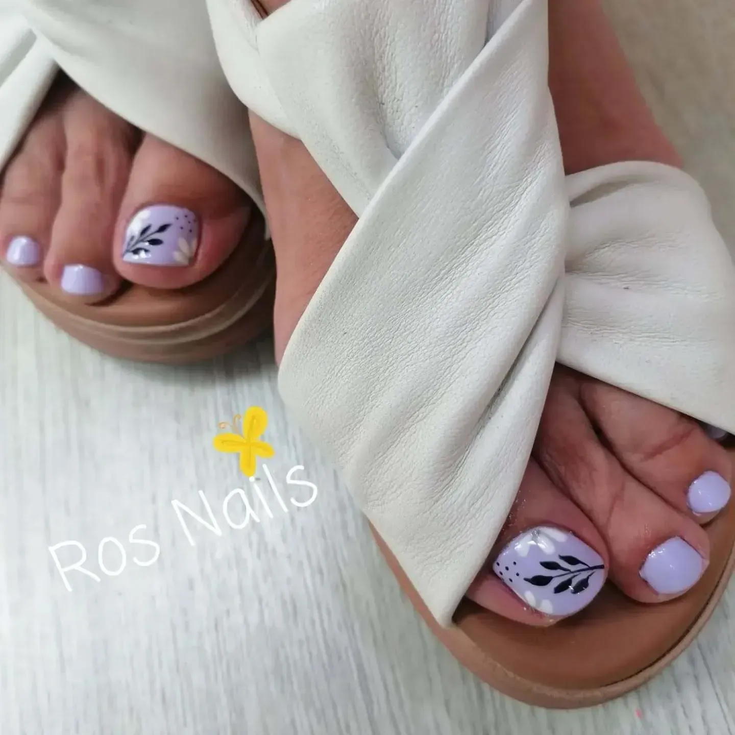 Pies en sandalias blancas con uñas color lavanda, una de ellas acentuada con un diseño de hojas negras.