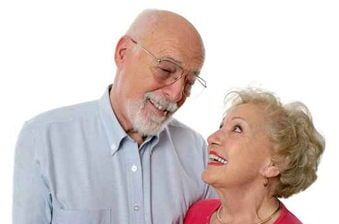 Casal de idosos sorridentes olhando um para o outro; homem de camisa azul clara, mulher de blusa vermelha.