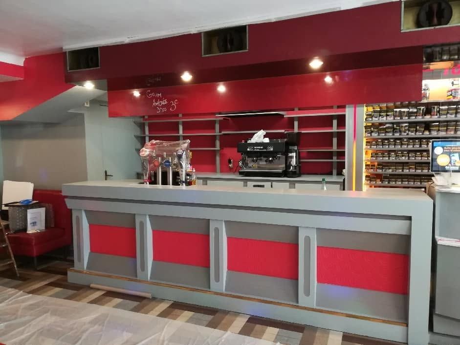 Comptoir de bar rouge et gris vide dans un salon, avec étagères et robinets derrière.