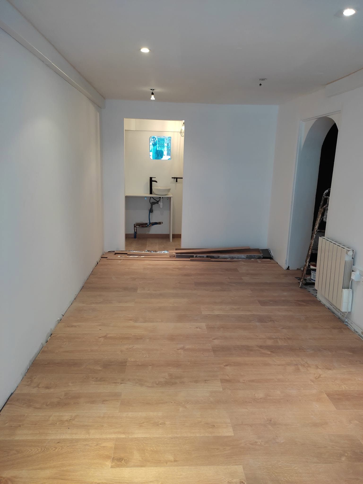 Pièce blanche vide avec parquet, spots encastrés et une porte donnant sur un petit coin lavabo.
