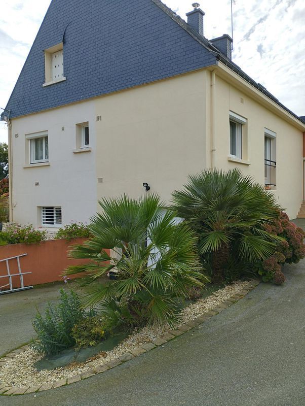 Maison au toit en ardoise pentu et aux murs couleur crème, bordée d'une allée paysagée ornée d'arbustes.