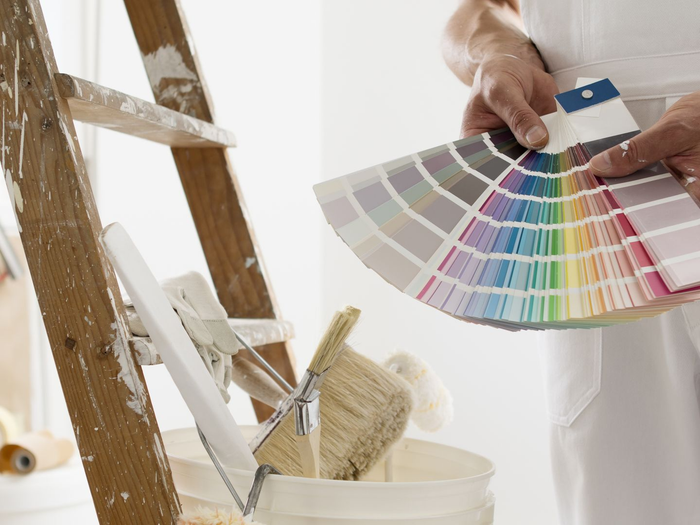 Une personne tient un éventail d'échantillons de couleurs près d'une échelle et d'un seau remplis de peinture.