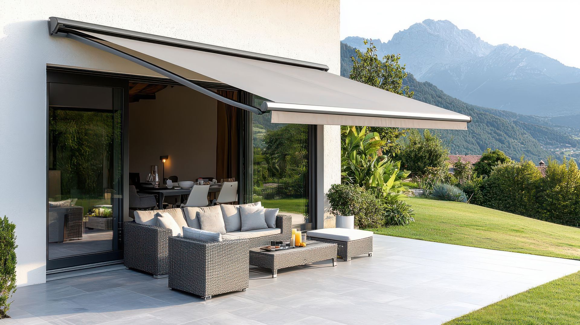 Patio moderne avec sièges en osier, auvent rétractable et vue sur la montagne à côté d'une maison aux murs de verre.