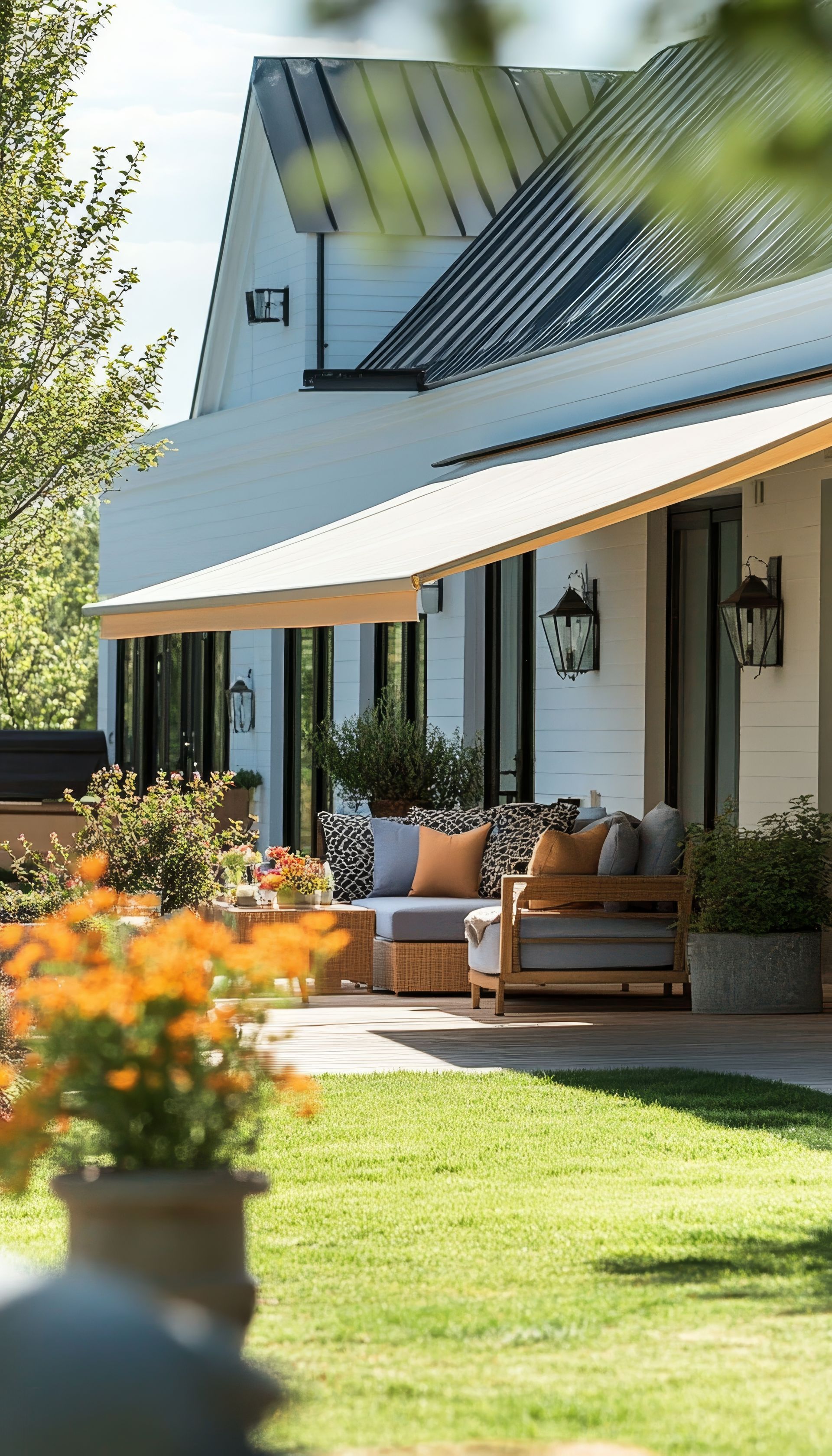 Maison moderne avec terrasse aménagée, grand auvent et pelouse verdoyante ensoleillée avec des fleurs orange au premier plan.