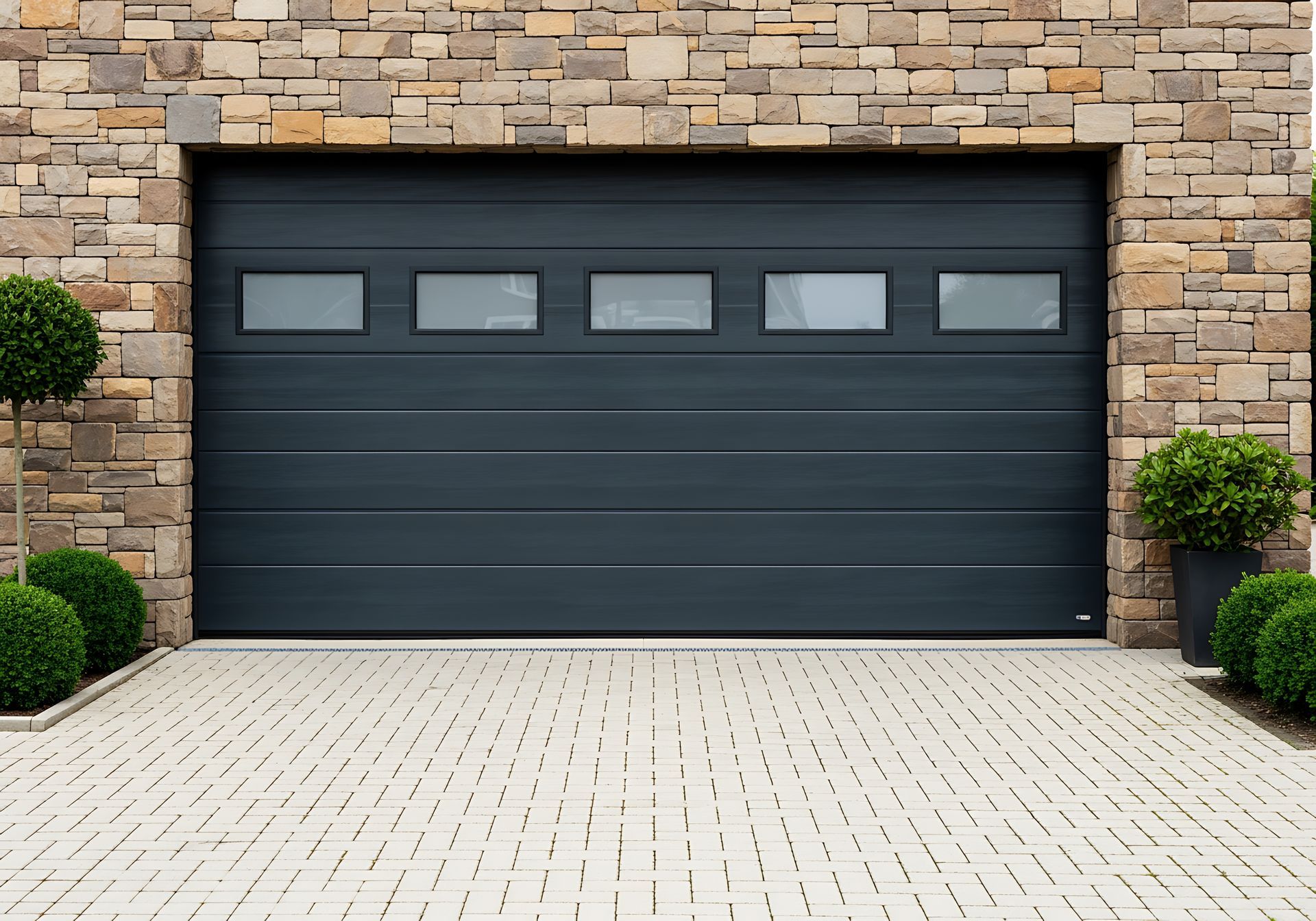 Porte de garage gris foncé intégrée à une façade de maison en pierre avec allée pavée et arbustes.