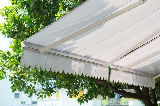 Auvent extérieur blanc à bordure festonnée, près d'arbres verts et d'un bâtiment.