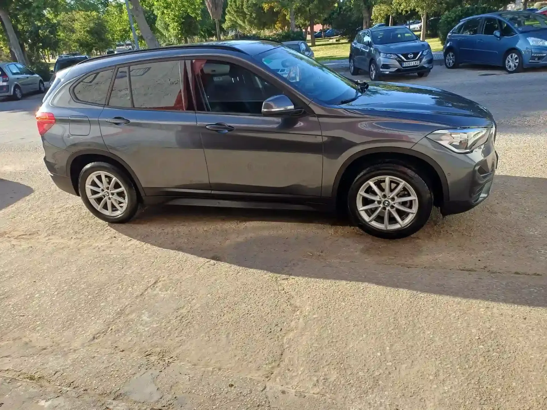Un SUV gris estacionado en una calle soleada con árboles y otros autos al fondo.