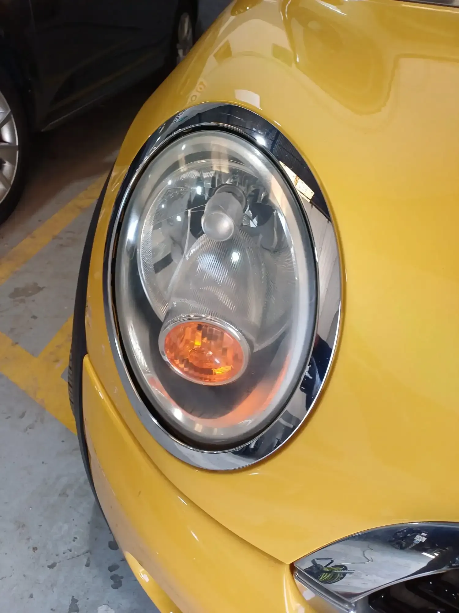 Primer plano del faro redondo de un coche amarillo con embellecedor cromado y luz intermitente ámbar.