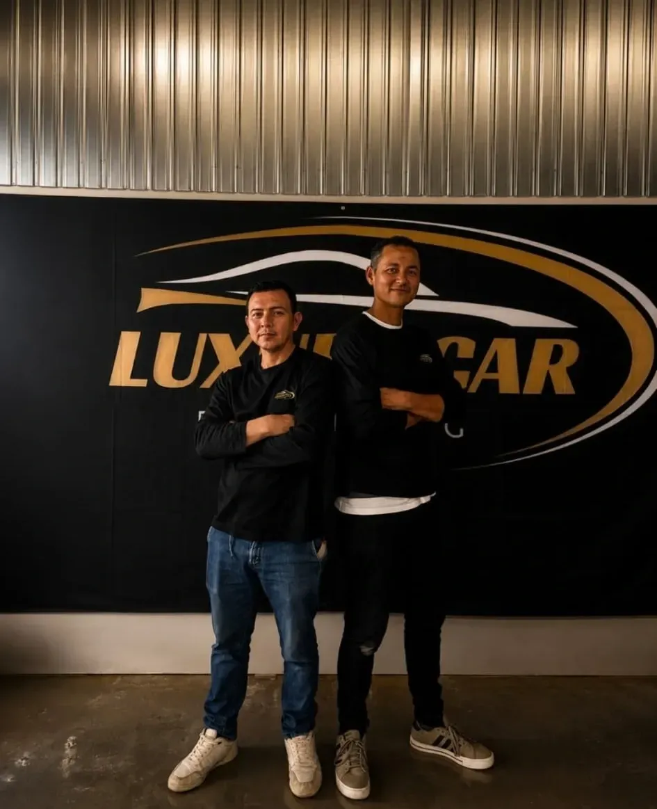 Dos personas posando con los brazos cruzados frente a una pared con el logotipo de Luxcar.