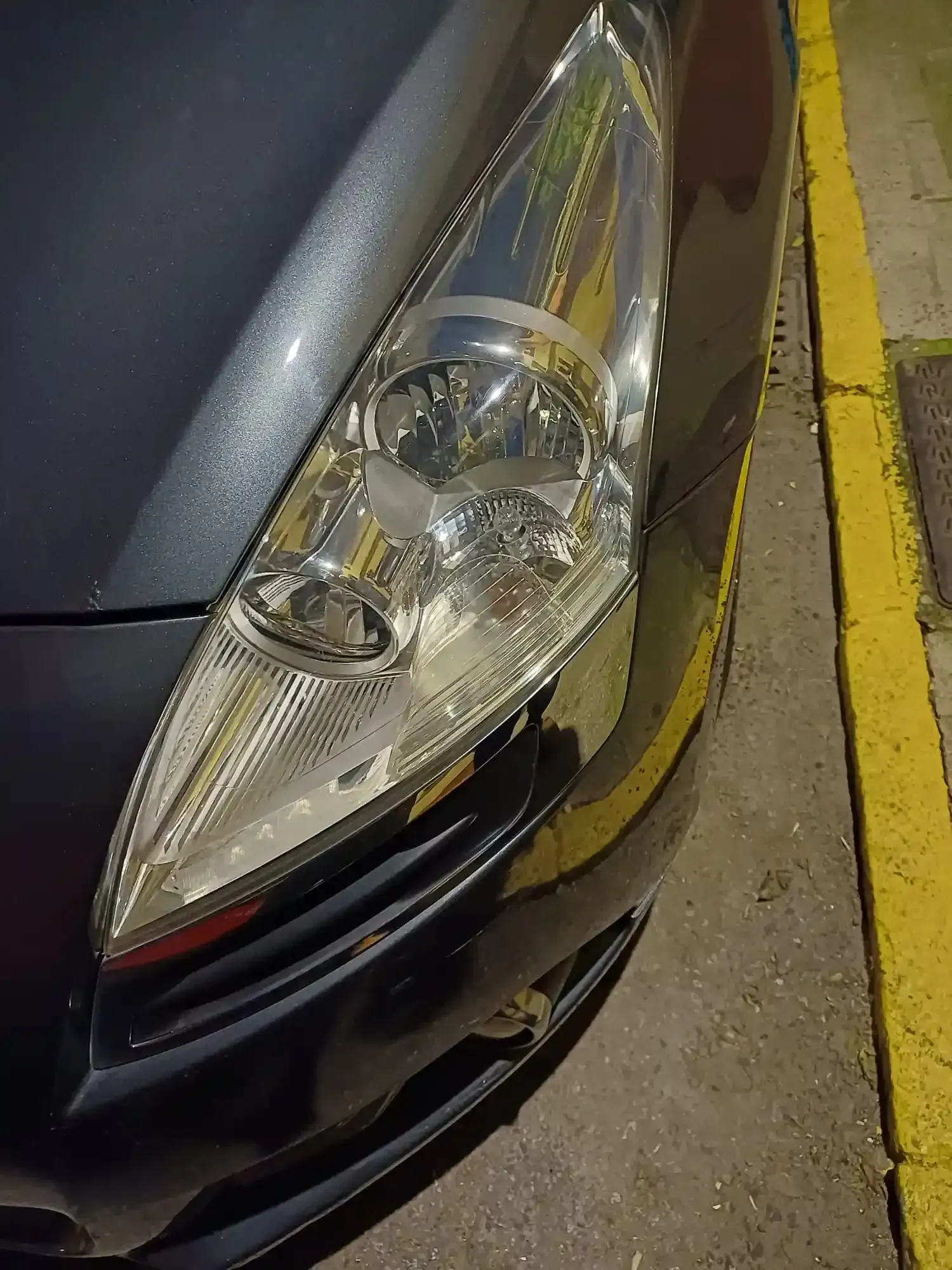 Primer plano del faro delantero y el parachoques de un coche aparcado junto a una línea amarilla en el bordillo.