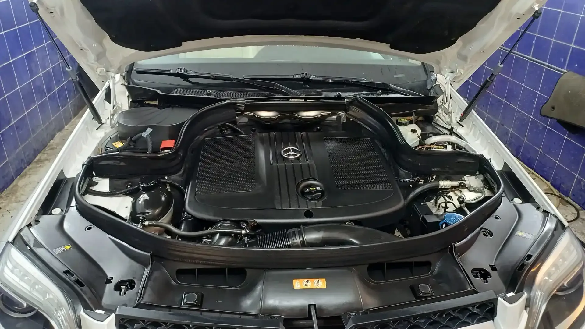 Capó abierto que deja ver el compartimento del motor con la cubierta negra en un garaje con azulejos azules.