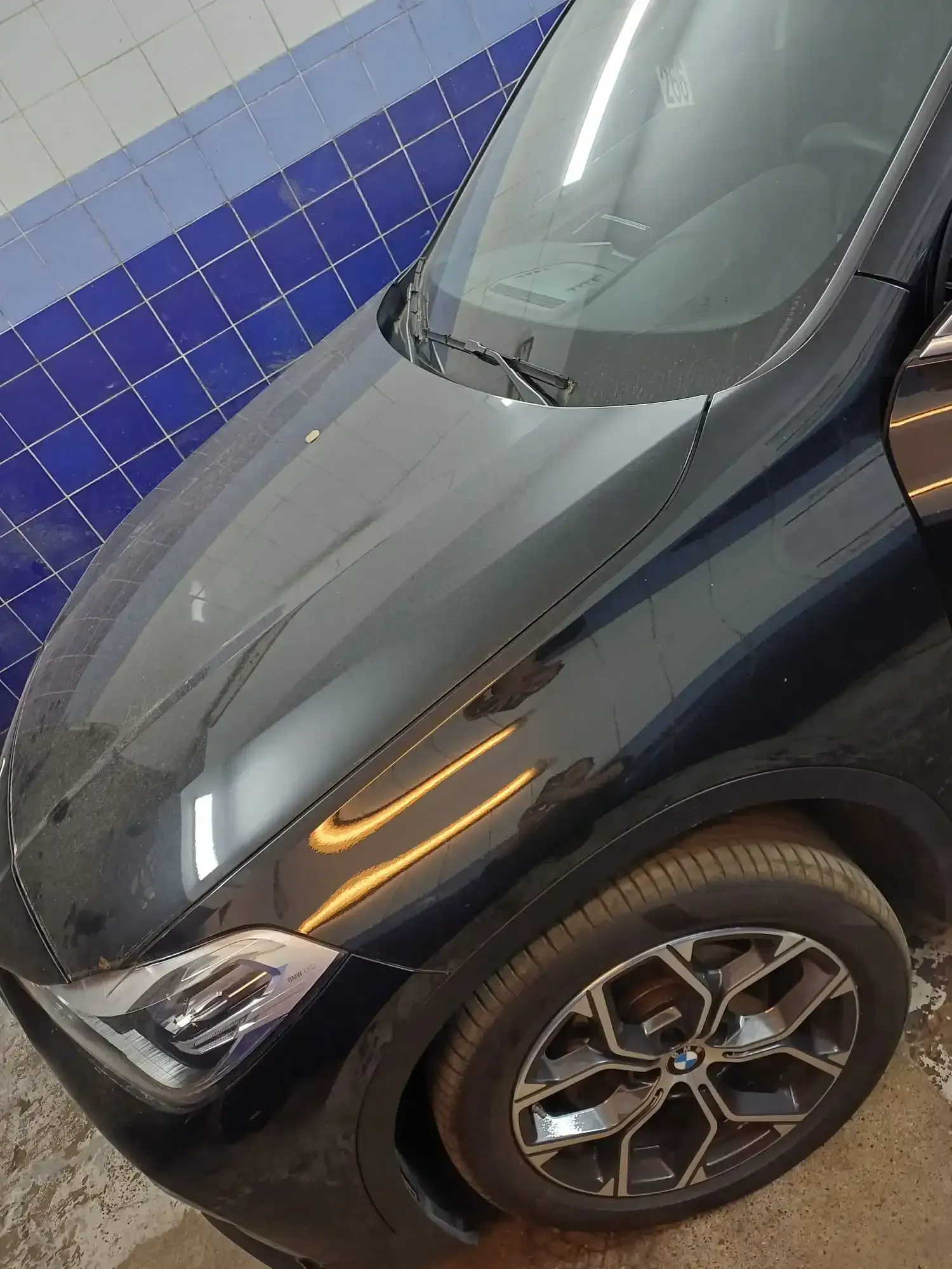 Coche negro aparcado junto a una pared de azulejos azules, visto desde el lado frontal izquierdo.
