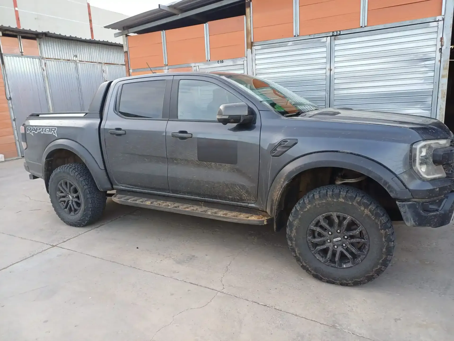 Camioneta de color gris oscuro estacionada en una entrada de vehículos junto a una cerca de metal y un edificio naranja.