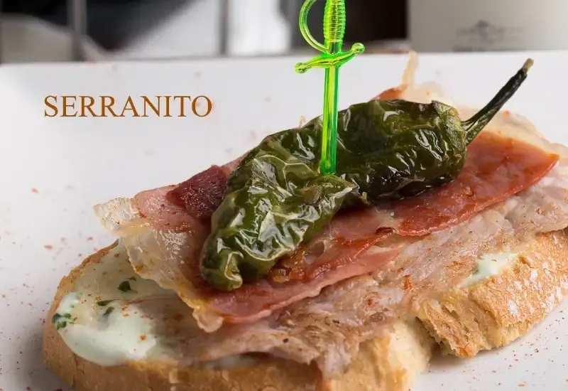 Sándwich abierto con pimientos asados ​​y jamón en pan tostado, etiquetado como “SERRANITO”.