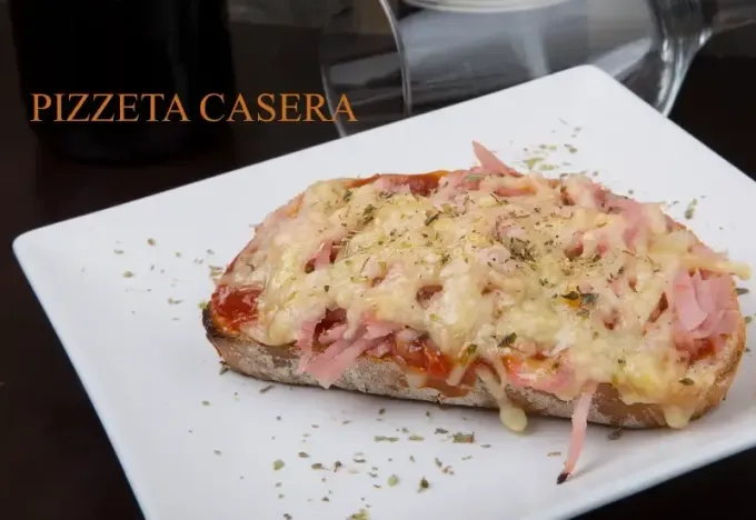 Rebanada de pizza casera en un plato blanco con queso fundido y jamón, etiquetada como “Pizzeta casera”.