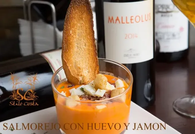 Salmorejo con huevo y jamón en vaso, con una tostada como adorno junto a una botella de vino.