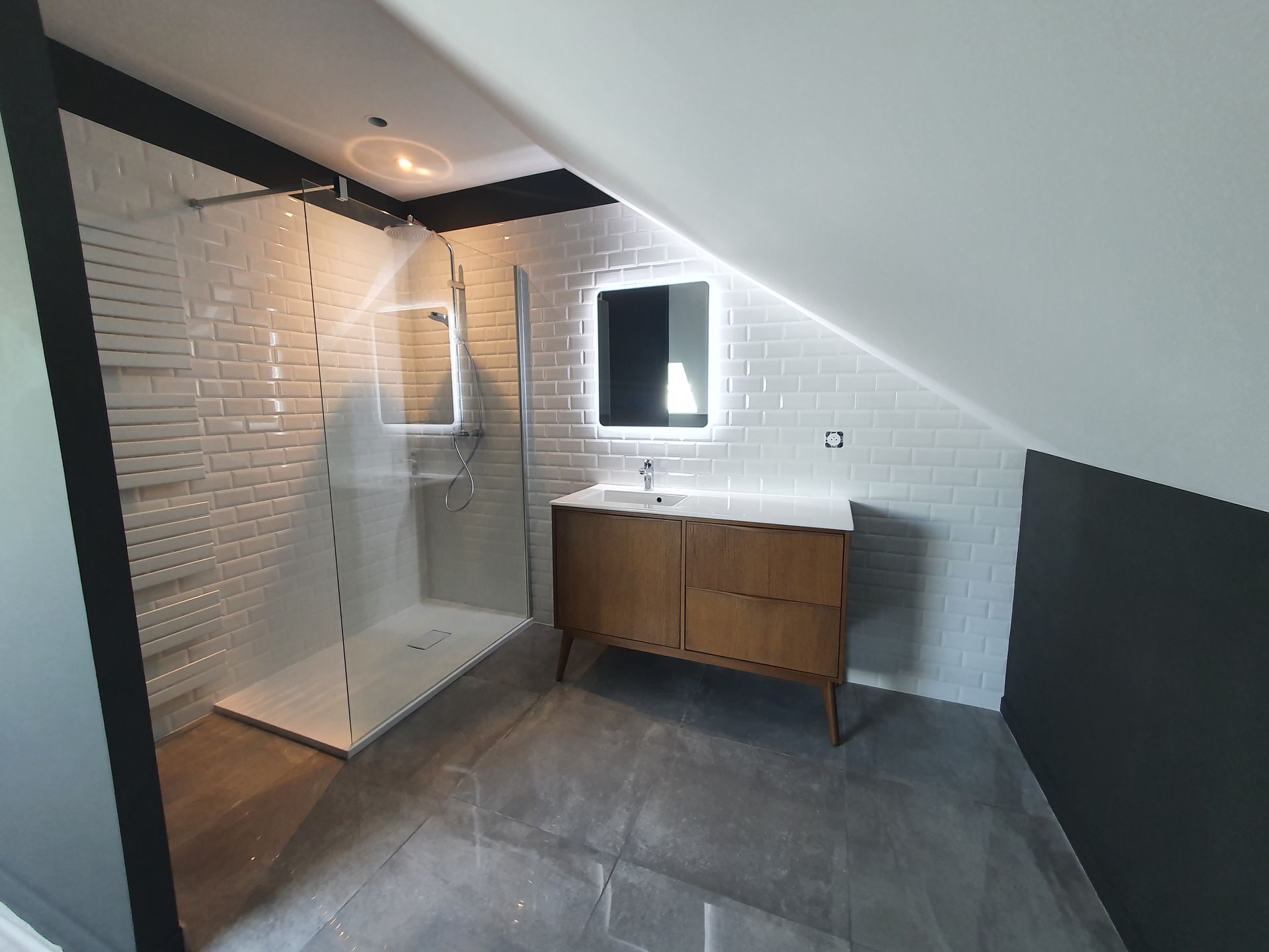 Salle de bains moderne avec douche carrelée blanche, meuble lavabo en bois, miroir et plafond mansardé