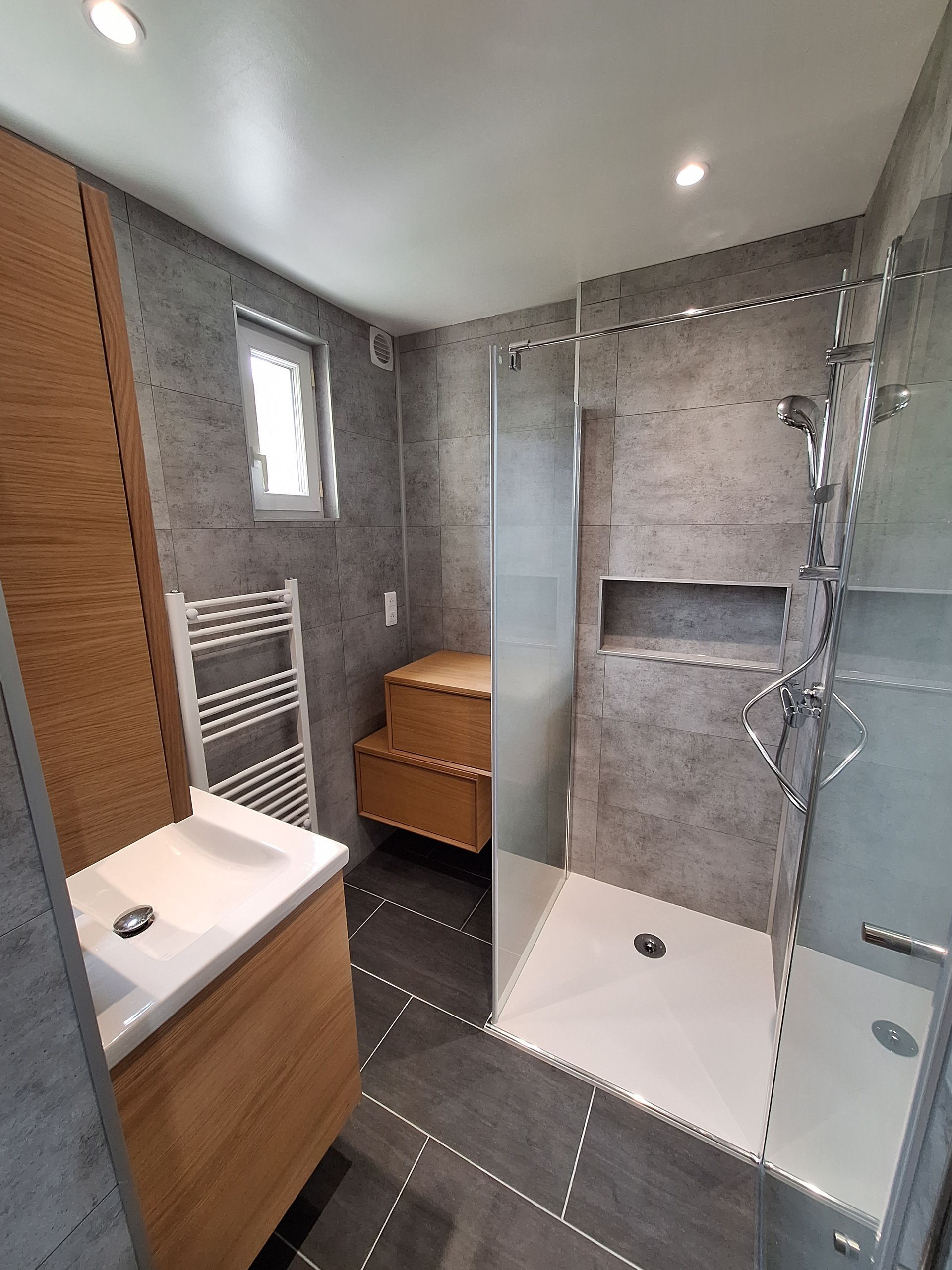 Salle de bains moderne avec douche à l'italienne, carrelage gris, lavabo blanc, sèche-serviettes et meuble lavabo en bois.