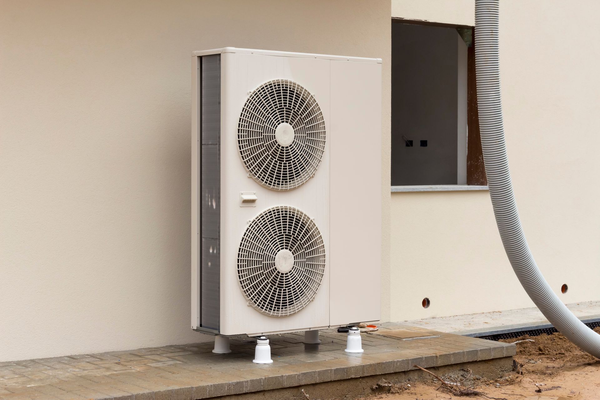 Unité de climatisation extérieure avec deux ventilateurs, adossée à un mur beige.