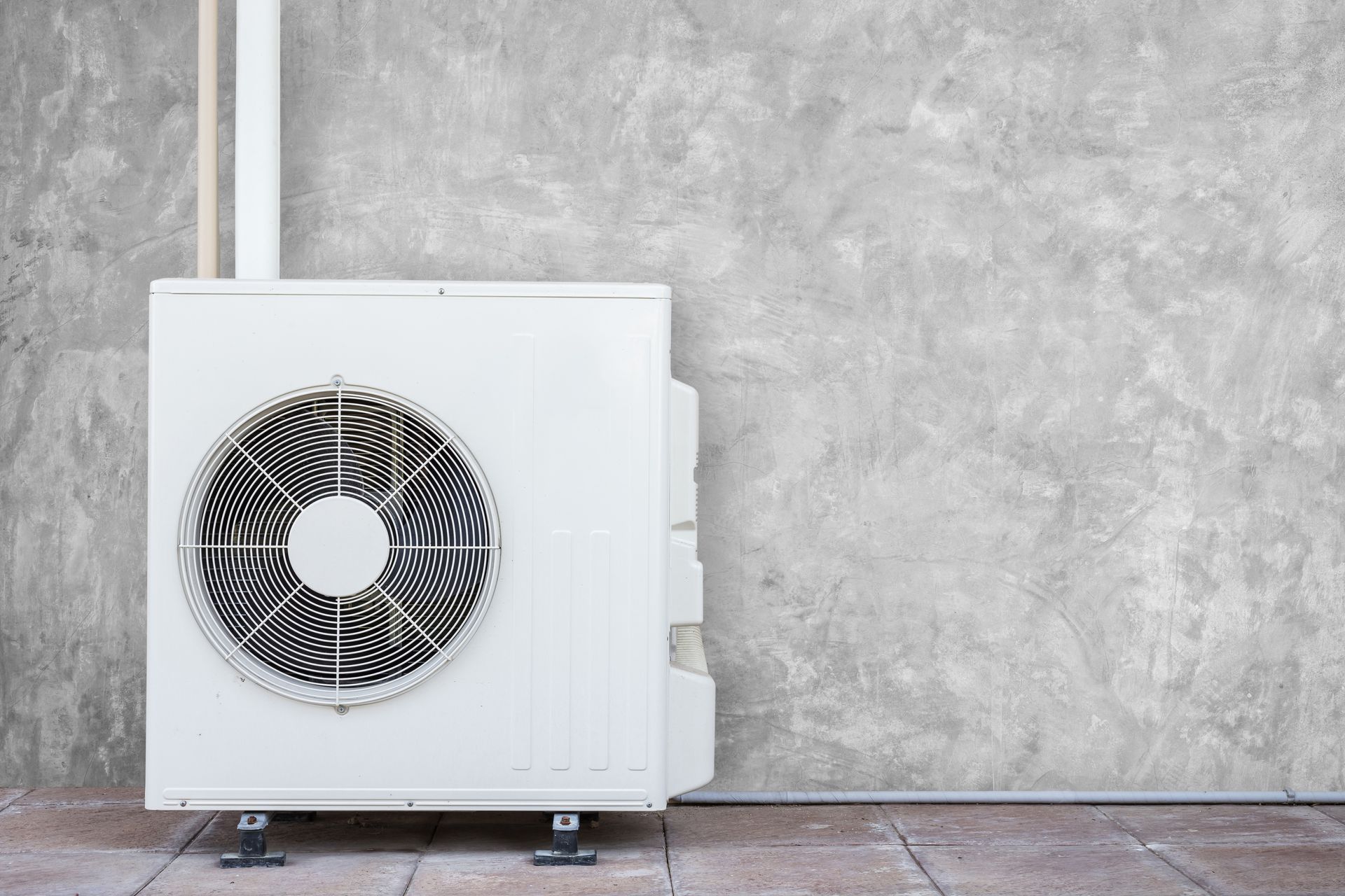 Unité de climatisation extérieure blanche sur un mur et un sol gris, avec un ventilateur frontal visible.