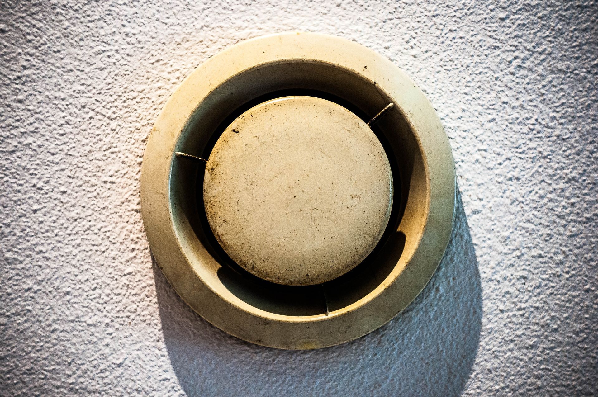 Grille d'aération murale ronde beige avec une ombre sur une surface blanche texturée.