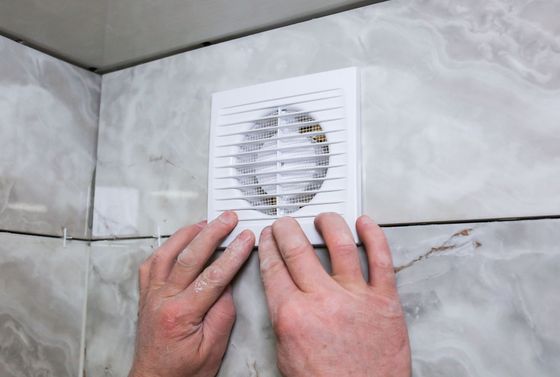 Des mains installent une grille d'aération blanche sur un mur carrelé gris.