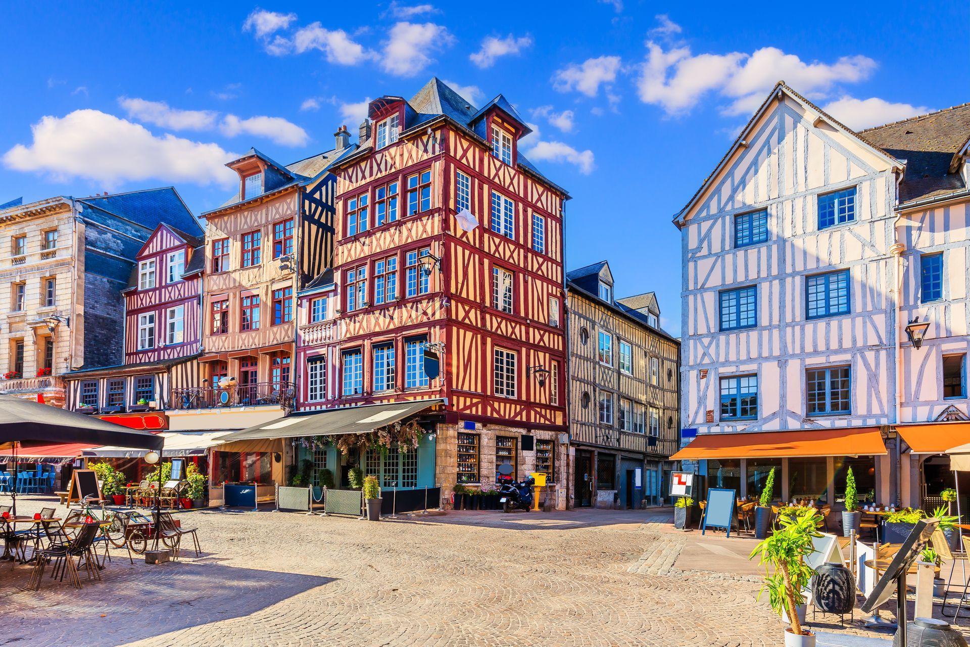 Des bâtiments colorés de style européen entourent une place pavée ensoleillée, bordée de cafés en terrasse, dans le centre-ville de Rouen.