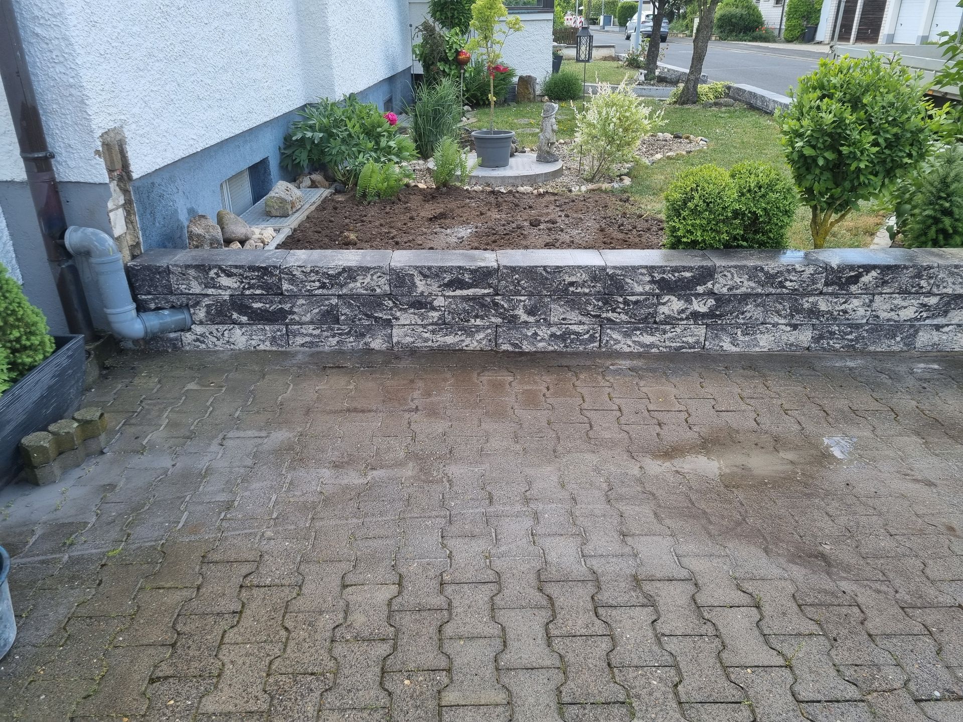 Eine mit Ziegeln gepflasterte Terrasse vor einem erhöhten Steinbeet mit Pflanzen, neben einem Gebäude mit einem Fallrohr.