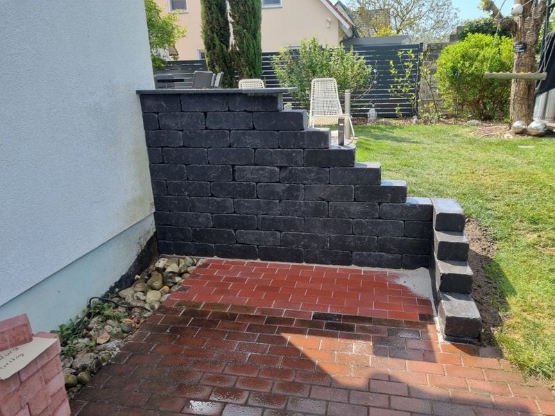 Eine Wohnstraße mit grauem Asphaltbelag und einer niedrigen Steinmauer, die an einen Rasen mit Büschen und Bäumen angrenzt.