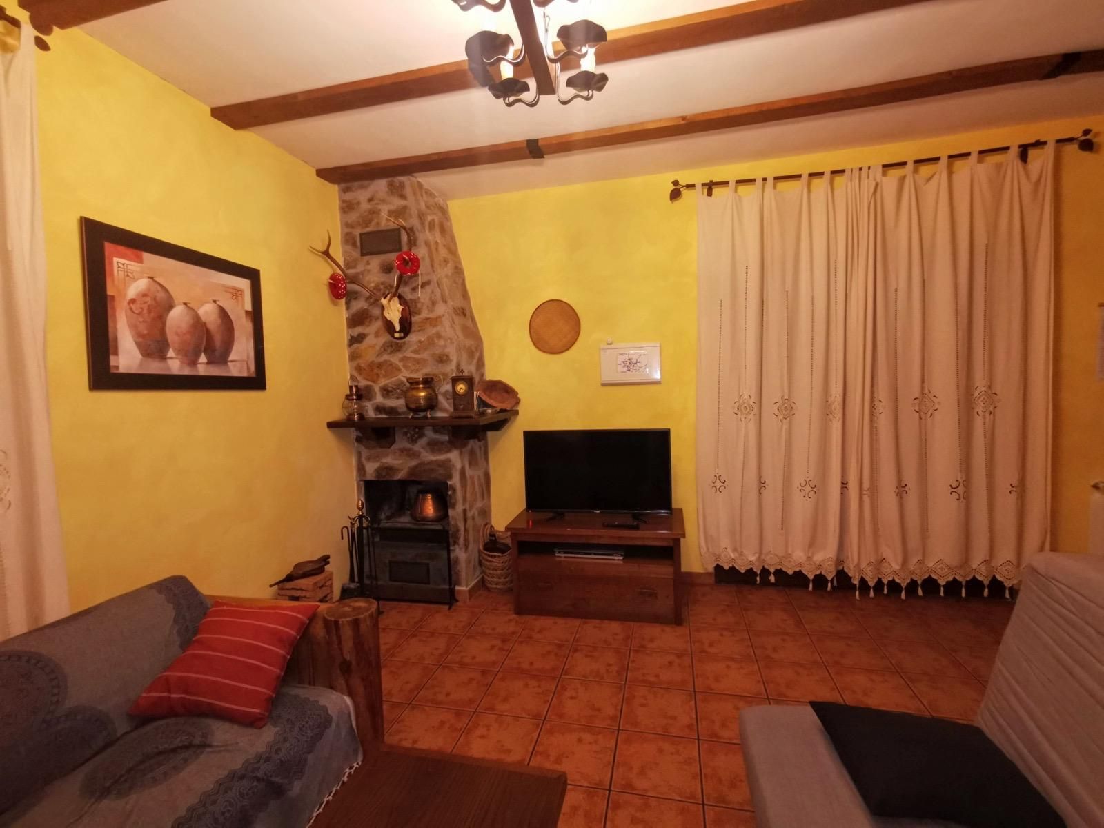 Acogedora sala de estar con paredes amarillas, chimenea, televisor, cortinas beige y sofás marrones sobre un suelo de baldosas.