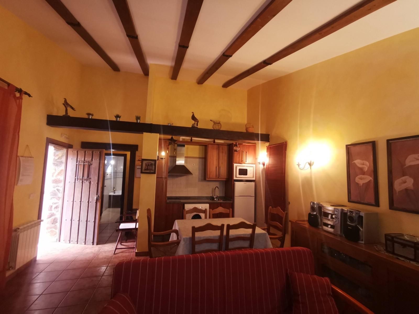 Acogedora sala de estar con sofá rojo, vigas de madera y una pequeña zona de cocina con luz cálida.