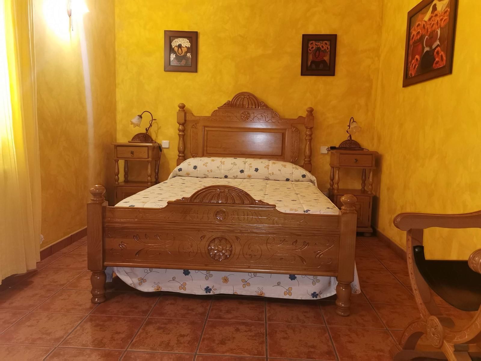 Acogedora habitación rústica con cama de madera, paredes amarillas, cuadros enmarcados y suelo de baldosas.