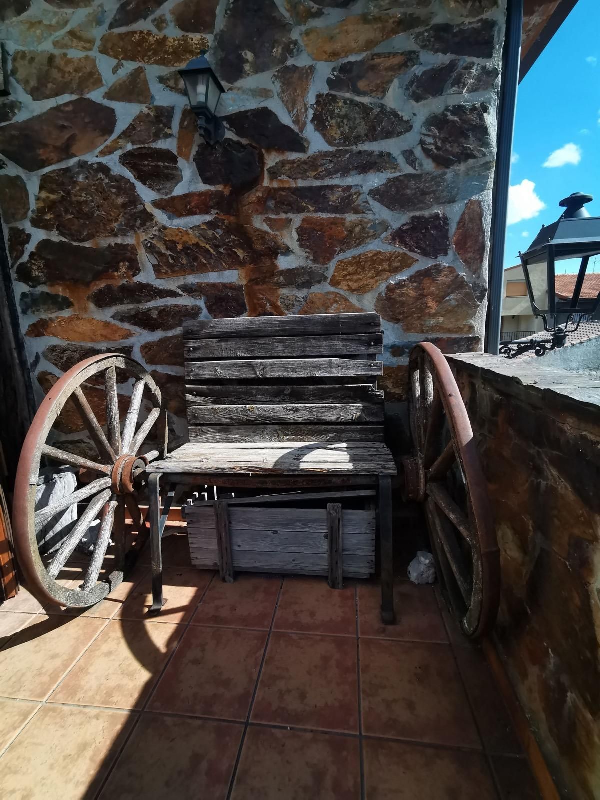 Banco rústico de madera y decoración con ruedas de carreta en un patio de piedra junto a un muro de piedra con vistas al mar.