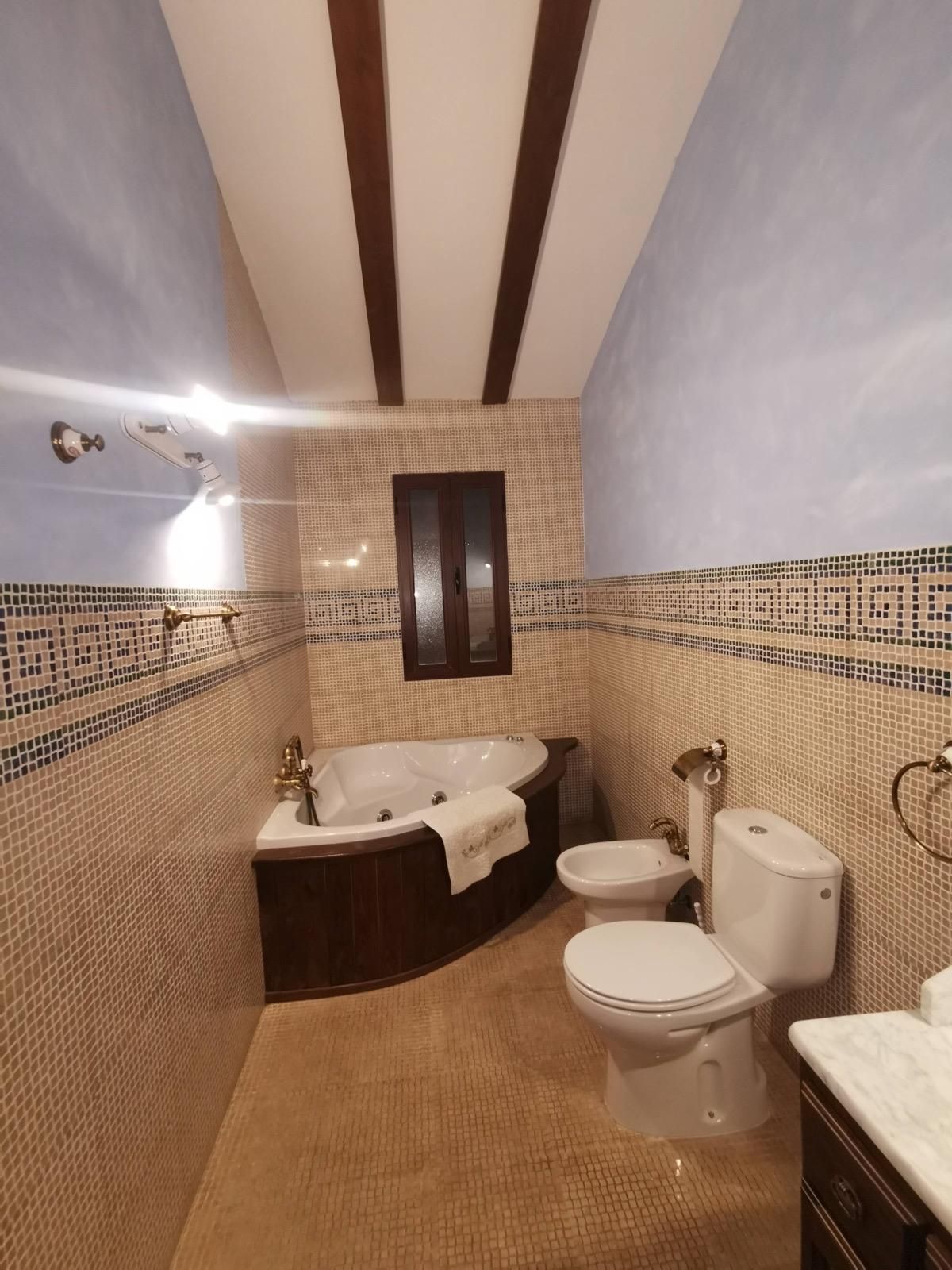 Pequeño baño alicatado con bañera de esquina, inodoro, bidé, lavabo y ventana esmerilada bajo vigas de techo a rayas.