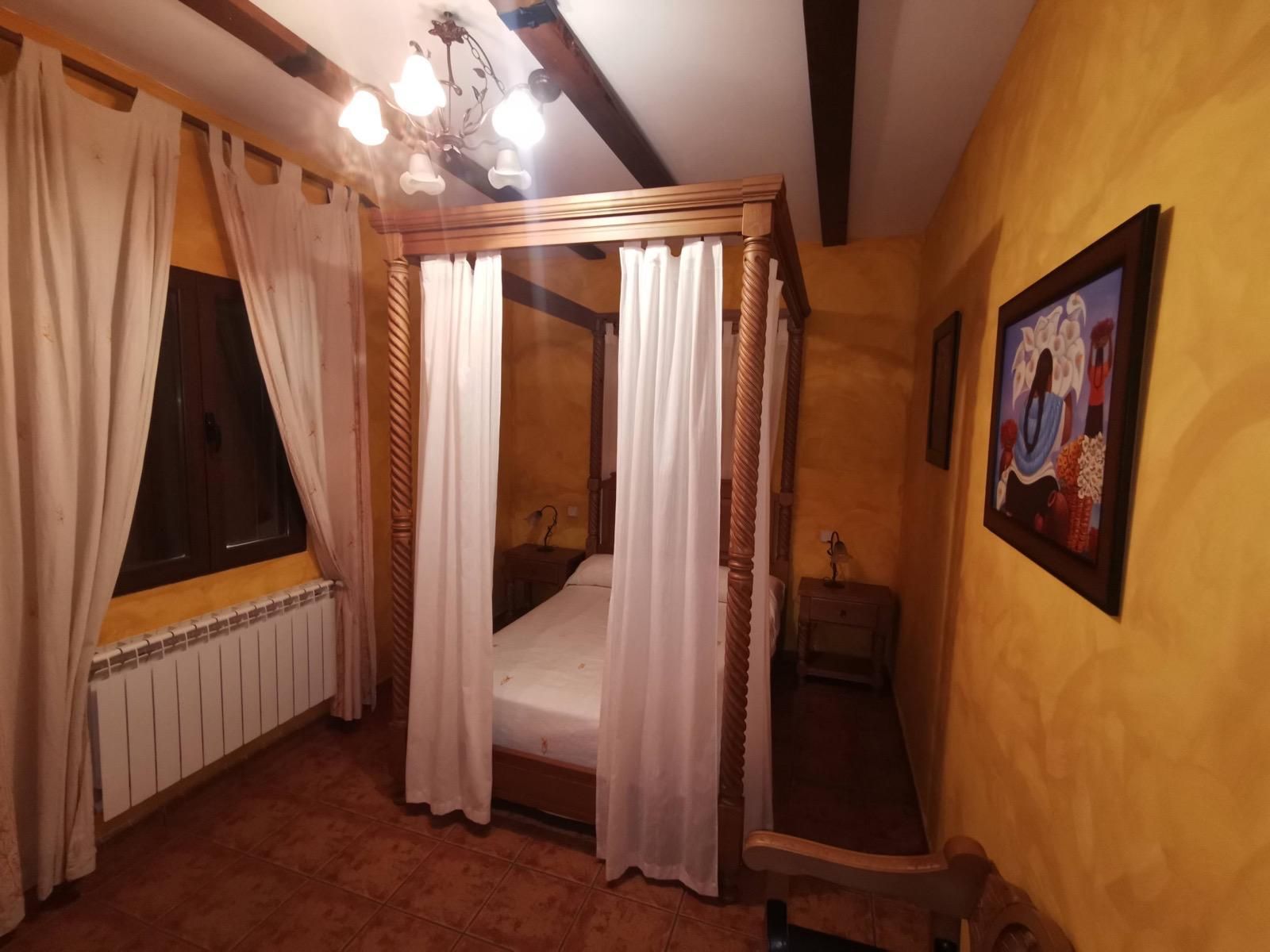 Acogedora habitación con cama con dosel, cortinas color crema, paredes de un cálido color amarillo y un cuadro enmarcado a la derecha.