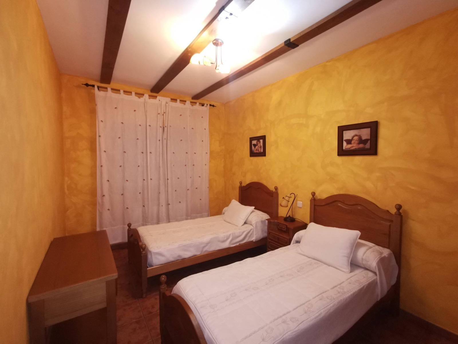 Habitación pequeña con dos camas individuales, muebles de madera y paredes de color amarillo cálido bajo una lámpara de techo.
