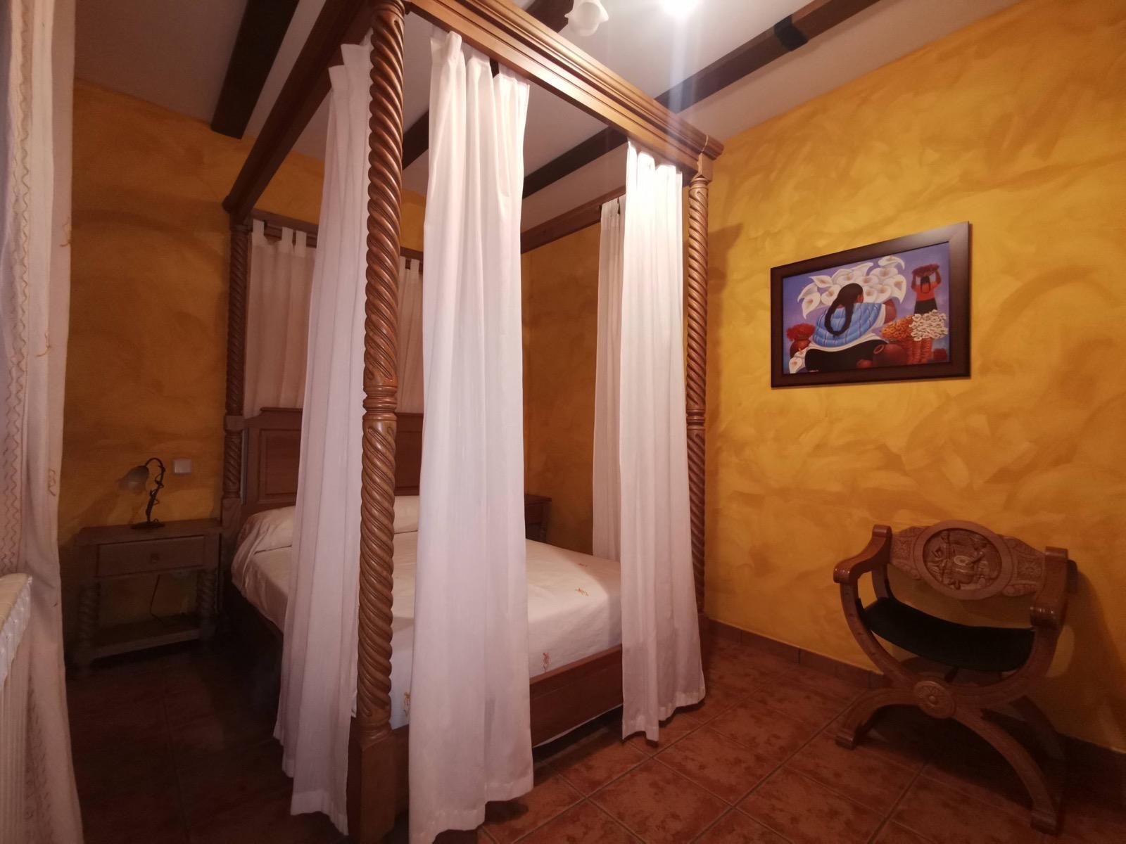Cama con dosel y cortinas blancas en un dormitorio de color naranja con iluminación cálida, decorado con cuadros en la pared y una silla de madera.