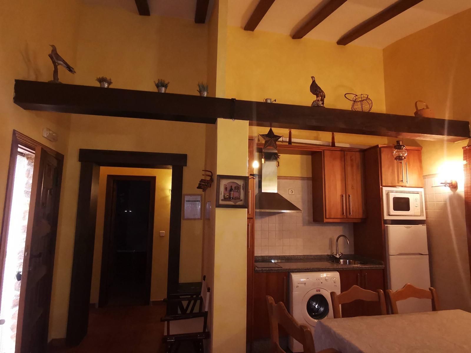 Acogedora cocina y comedor con vigas de madera, iluminación cálida, refrigerador, estufa y lavadora.