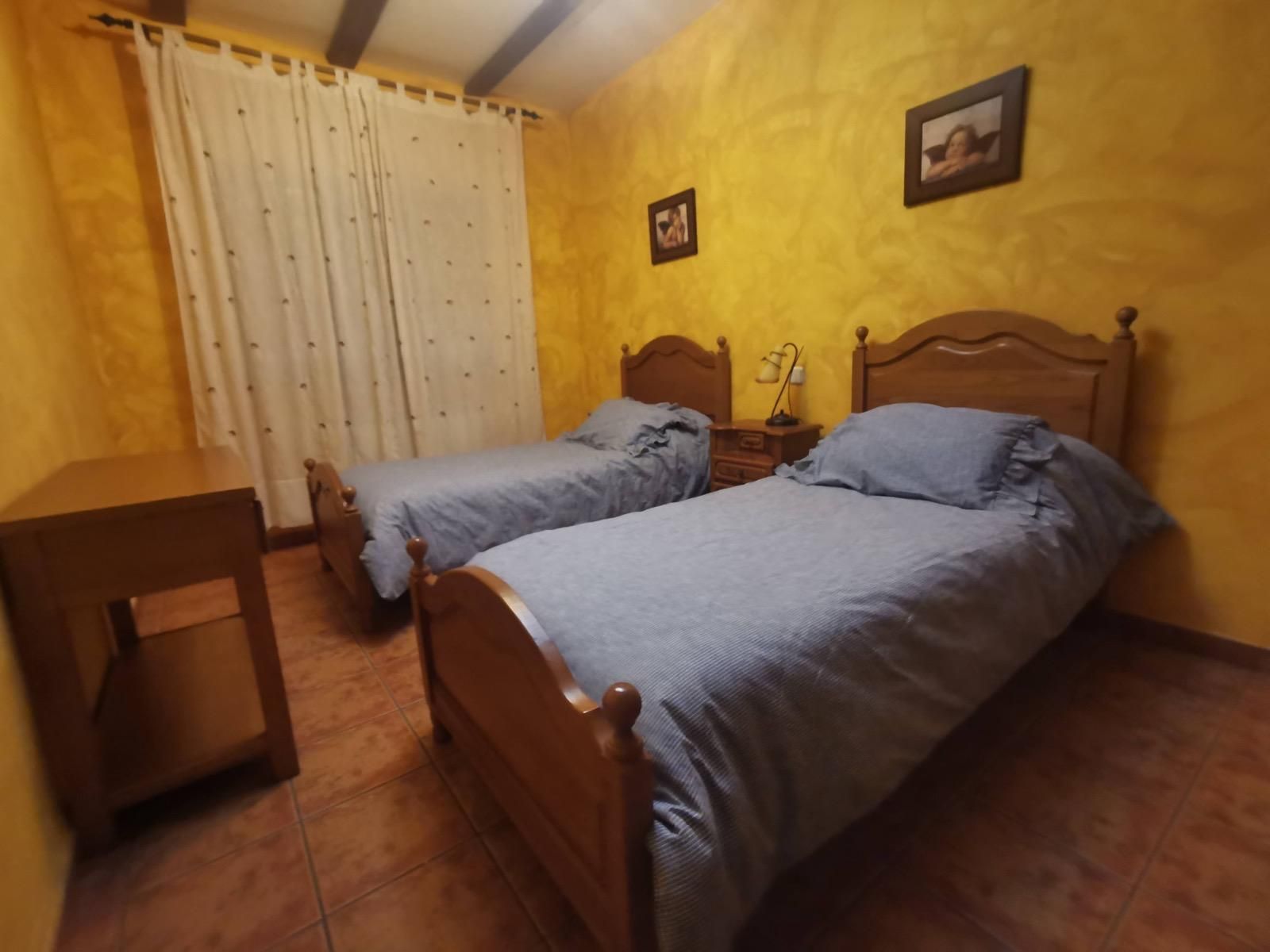 Habitación en tonos cálidos con dos camas individuales de madera, paredes amarillas y una ventana con cortinas.