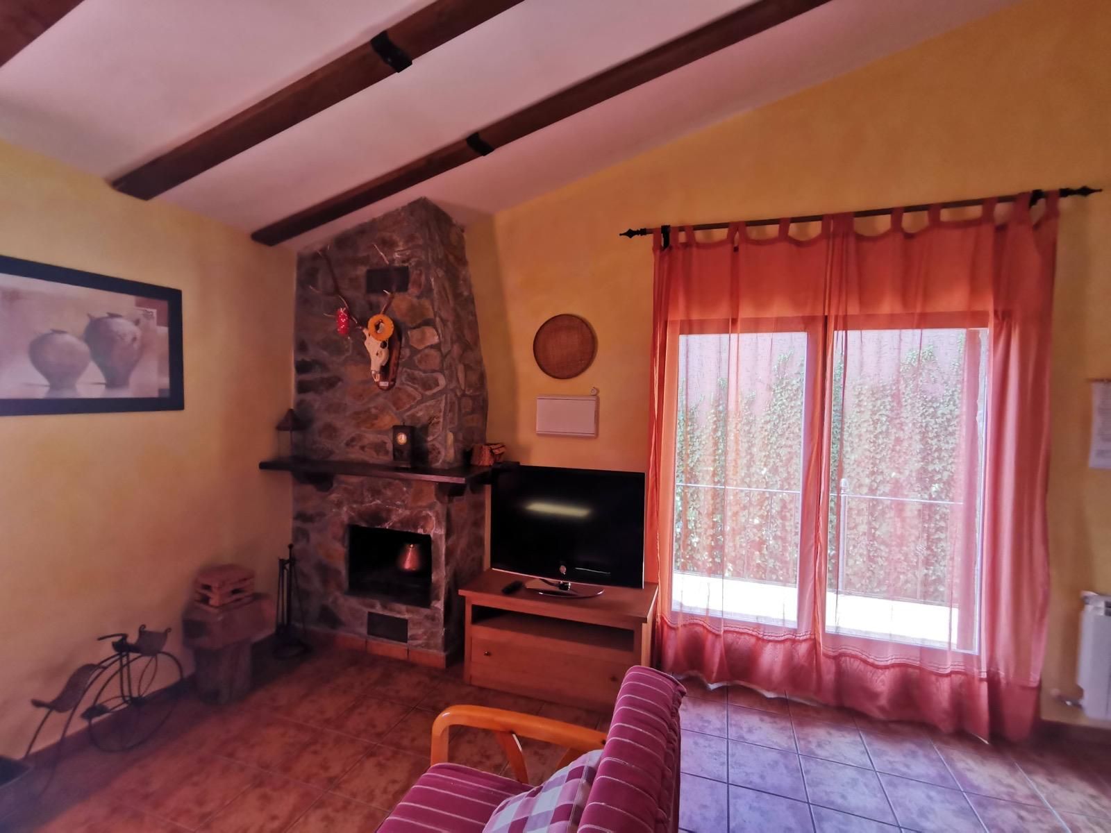 Acogedora sala de estar con chimenea de piedra, televisor, cortinas rojas y techo inclinado con vigas de madera.