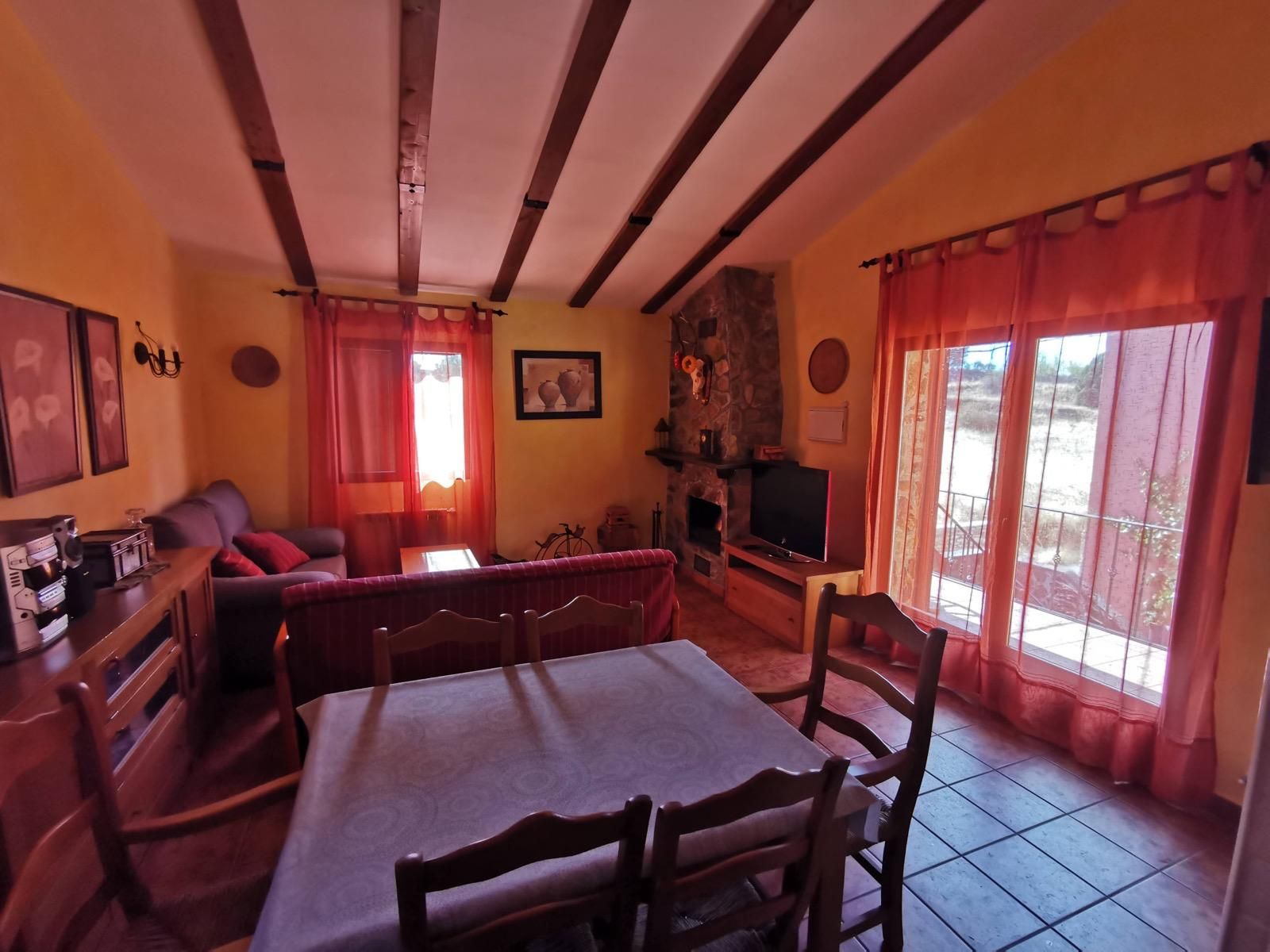 Acogedora sala de estar con cortinas rojas, vigas de madera, chimenea y un sofá frente al televisor.