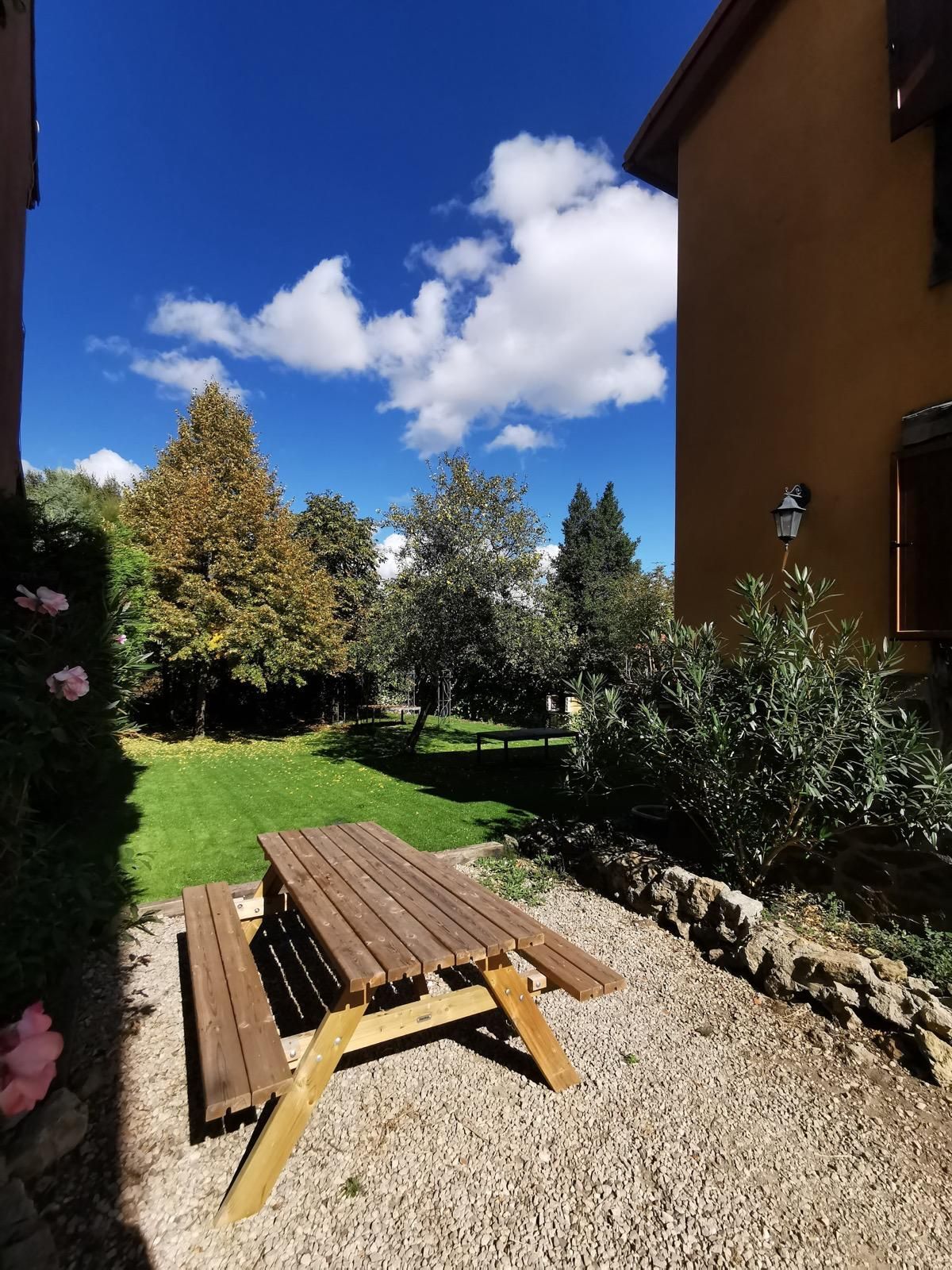 Patio trasero con mesa de picnic de madera, suelo de grava, césped verde, árboles y cielo azul con nubes.