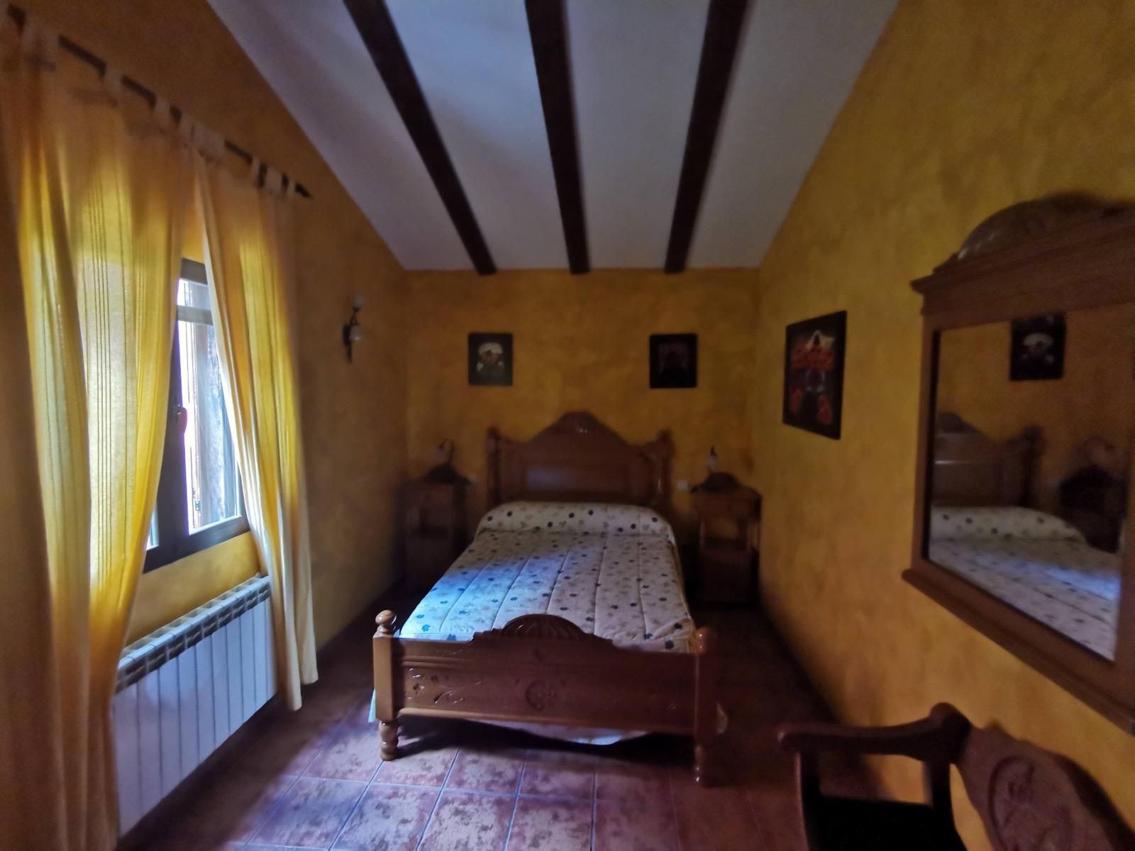 Acogedora habitación con paredes amarillas, cama de madera, cortinas a rayas, espejo y vigas en el techo.