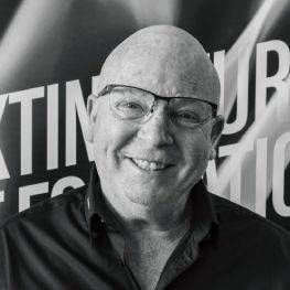 Portrait en noir et blanc d'un homme chauve souriant portant des lunettes devant un fond de marque