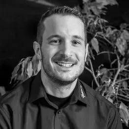 Un homme souriant en chemise sombre pose à l'intérieur, à côté d'une plante à feuillage noir et blanc.