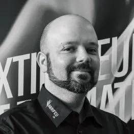 Un homme chauve et barbu, souriant et vêtu d'une chemise sombre, se tient devant un fond graphique flou.