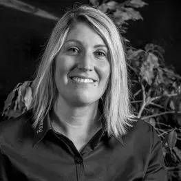 Femme souriante en chemise sombre sur fond d'intérieur flou.